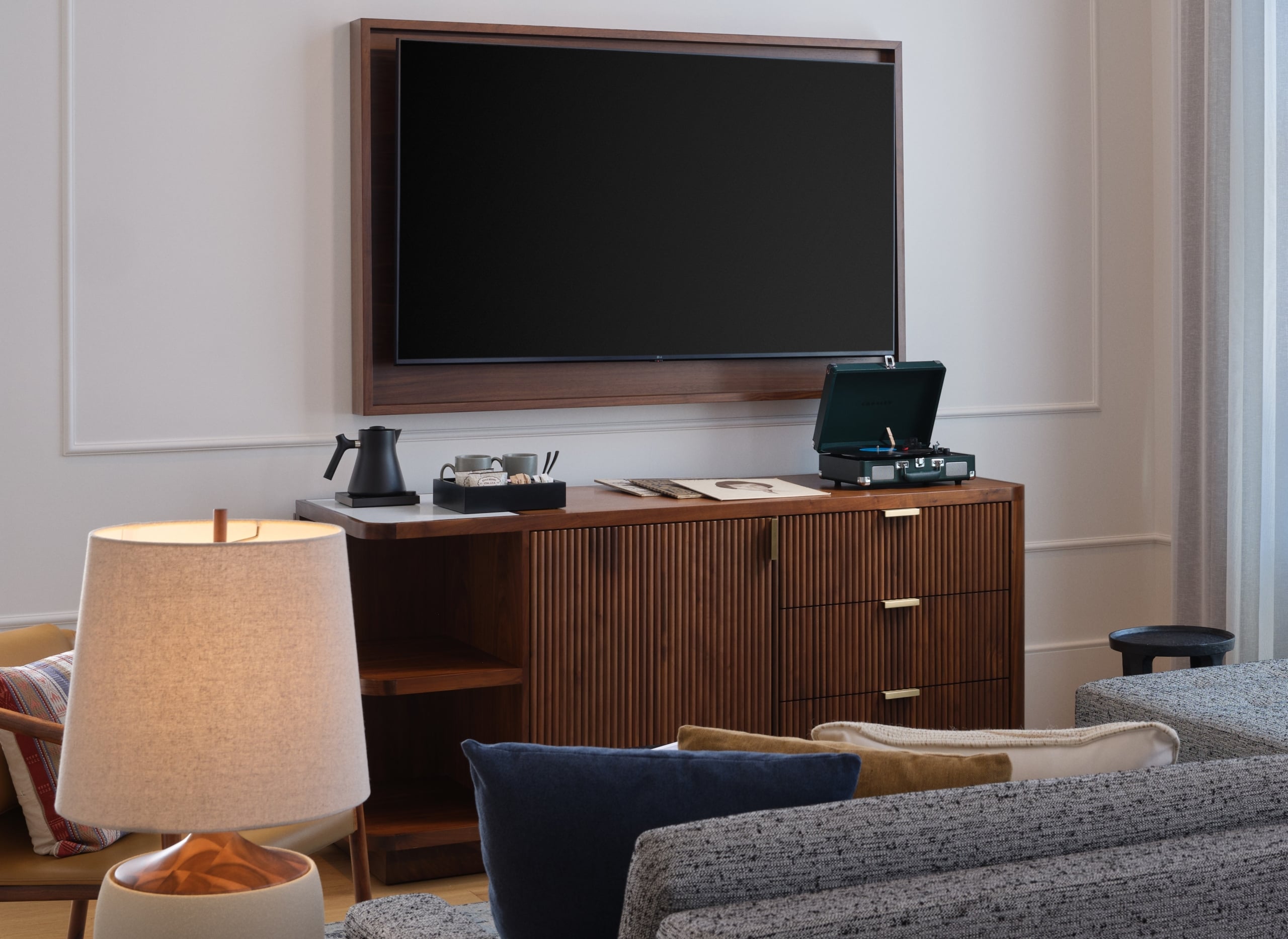 A wooden dresser sits against a white wall with a TV hung above it and a coffee maker and record player on top of it. A sectional couch faces the TV.